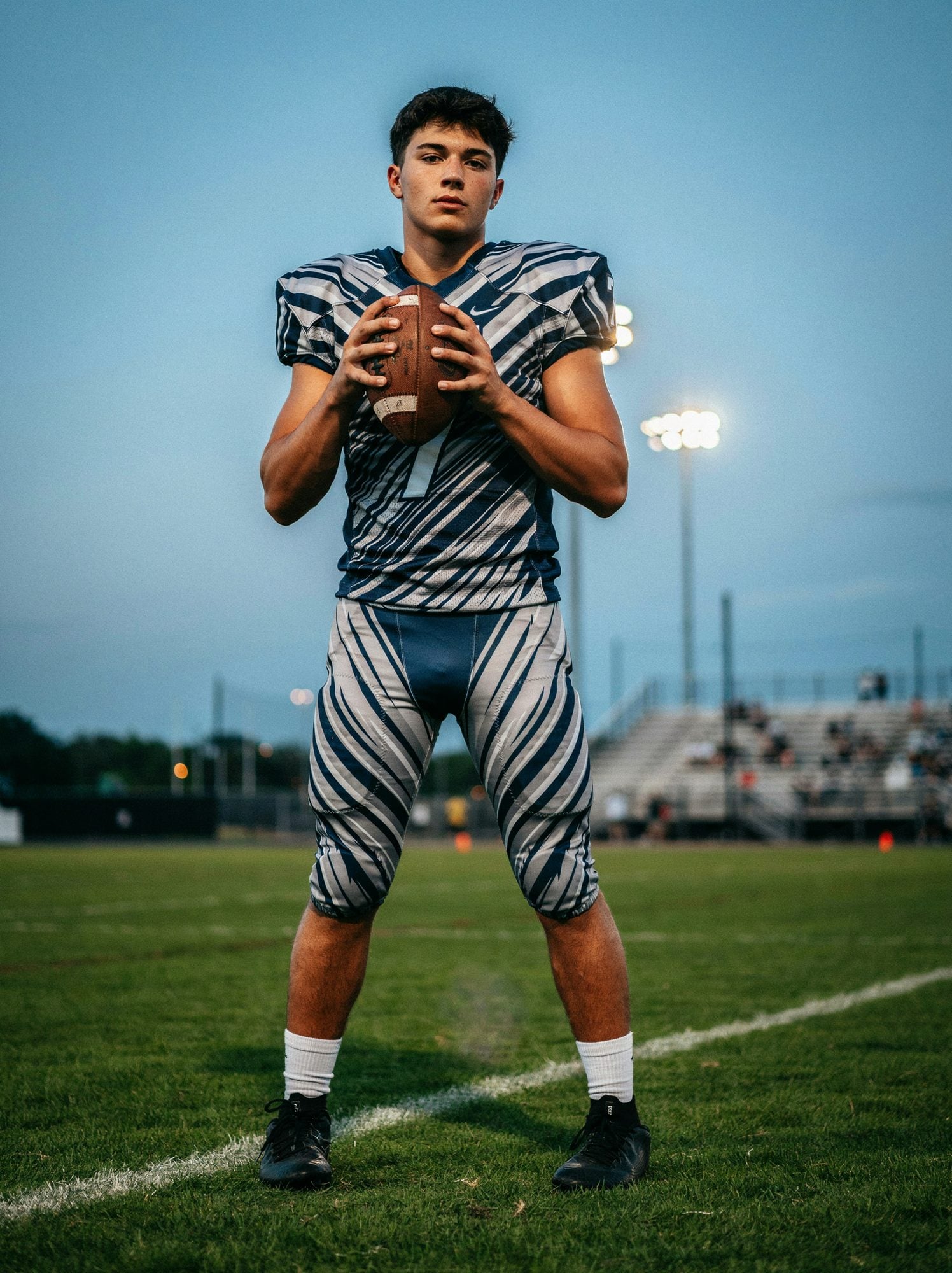 A quarterback holding a football in a custom navy and silver striped uniform under stadium lights